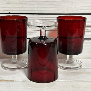 Pode incluir: Três taças de vinho vermelho rubi com hastes e bases transparentes. Uma taça está de cabeça para baixo, mostrando a base transparente. As outras duas taças estão em pé, exibindo a cor rica do vidro. As taças estão sobre um fundo de madeira clara.