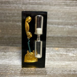 May include: A black rectangular decorative piece featuring a gold-colored vintage telephone and a clear hourglass with white sand. The telephone has a coiled green cord. The hourglass is silver-toned.