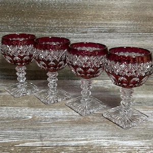 Pode incluir: Um conjunto de quatro taças de vinho com bojos vermelho-escuro e um padrão de diamante transparente. Cada taça tem uma haste transparente multifacetada e uma base quadrada com um design de raios de sol, mostrando uma transição clara.
