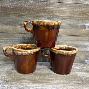 May include: Three brown ceramic mugs with a drip glaze design. The mugs have a rounded shape with a handle and a lighter-colored rim. The mugs are stacked on top of each other.