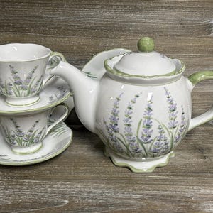 May include: A white ceramic tea set featuring a lavender floral pattern with green accents. The set includes a teapot with a lid and two teacups with matching saucers. Each piece has a scalloped edge and a painted green rim detail.