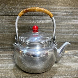 Könnte beinhalten: Eine silberne Vintage-Teekanne mit einem geflochtenen Griff und einem roten Knopf auf dem Deckel. Die Kanne hat einen Ausguss zum Ausgießen und ist aus glänzendem Metall gefertigt. Der Griff ist gebogen und am Körper der Kanne befestigt.