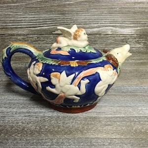 May include: A blue and white ceramic teapot with a cherub design. The teapot has a spout shaped like a dolphin and a lid with a cherub on top. The teapot is decorated with cherubs and flowers.