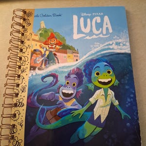 May include: A spiral-bound notebook featuring the Disney Pixar Luca Little Golden Book cover art. The front shows two sea creatures swimming in blue water, with a character on a bicycle in the background. The spine has a gold-colored metal coil.