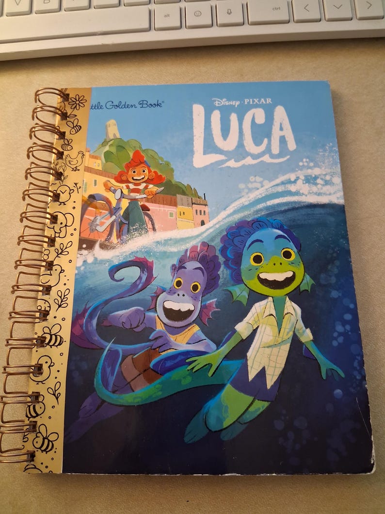 May include: A spiral-bound notebook featuring the Disney Pixar Luca Little Golden Book cover art. The front shows two sea creatures swimming in blue water, with a character on a bicycle in the background. The spine has a gold-colored metal coil.