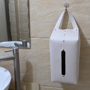 May include: White tissue box holder with a flap opening and a black opening for tissues. The holder is hanging on a wall with a hook.