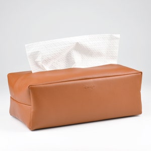 May include: A rectangular, brown tissue box with a simple design. White tissues are visible, emerging from the top opening. The box has a smooth, leather-like texture and a minimalist aesthetic. The brand name is subtly embossed on the front.