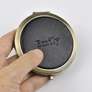 May include: Circular, antique brass compact mirror with a black, textured centre. The name "Emily" is embossed on the centre. The compact is held in a hand against a white background.