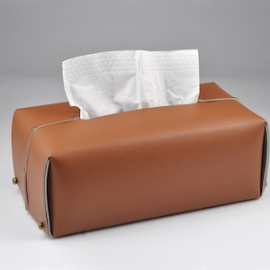 May include: A rectangular tissue box cover in a smooth tan finish with light-coloured edge piping and small metallic studs at the corners. A white tissue is pulled through the top opening, extending upwards from the centre of the box.