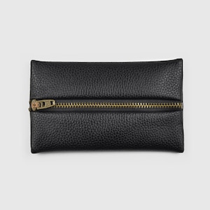 Könnte beinhalten: Eine rechteckige schwarze Tasche mit genarbter Textur und einem horizontalen, messingfarbenen Metallreißverschluss in der Mitte. Der Reißverschlussanhänger befindet sich auf der linken Seite der flachen und strukturierten Tasche.