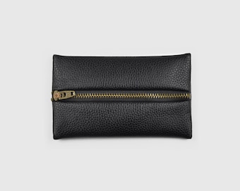 Può includere: Una pochette rettangolare nera con texture granulata, dotata di una chiusura a zip orizzontale in metallo color ottone al centro. Il cursore della zip è visibile sul lato sinistro della pochette dal design piatto e strutturato.