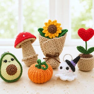 Op de afbeelding: Een verzameling van zes kleine gehaakte items, waaronder een avocado-sleutelhanger, een rood-witte paddenstoel, een zonnebloem in een beige wikkel, een oranje pompoen, een witte spook-sleutelhanger met een zwarte hoed en een rode hartvormige bloem.