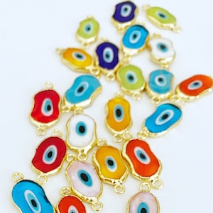 May include: A collection of colorful evil eye charms with gold accents. The charms are shaped like an eye and have a blue pupil. They are arranged in a pile on a white surface.
