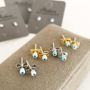 May include: A set of six pairs of small, silver stud earrings with blue and white evil eye charms. The earrings are displayed on a brown velvet cushion.
