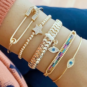 Pode incluir: Um conjunto de pulseiras de ouro com um fecho de segurança com uma estrela, uma pulseira de elos, uma pulseira de contas com um fecho de olho arco-íris e uma pulseira de ouro simples com um fecho de olho mau.