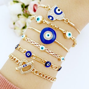 May include: A set of seven gold bracelets with blue evil eye charms. The bracelets feature different styles, including a chain link bracelet, a braided bracelet, and a bracelet with a heart-shaped charm. The charms are all decorated with blue enamel and white accents.