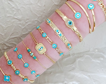 Può includere: Una collezione di braccialetti dorati con ciondoli blu a forma di occhio turco. I braccialetti sono disposti su un cuscino di velluto rosa.