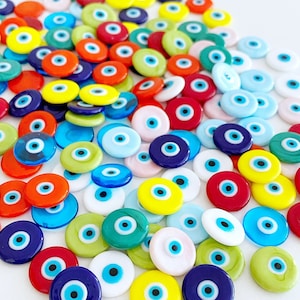 Könnte beinhalten: Eine Sammlung bunter böser Augen aus Glas, auch bekannt als Nazar Boncugu, in verschiedenen Farben wie Blau, Grün, Rot, Gelb und Weiß. Die Charms sind rund und haben ein blaues Augenmotiv in der Mitte.