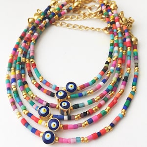 May include: Multicolored beaded bracelets with gold accents and blue evil eye charms. The bracelets are layered and feature a variety of colors, including pink, red, blue, green, and yellow.