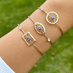 Op de afbeelding: Drie gouden armbanden met blauwe boze oog-charms. De armbanden zitten om de pols van een persoon. De charms hebben verschillende vormen, waaronder een cirkel, een ovaal en een vierkant.
