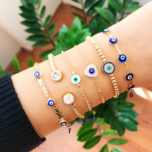 May include: Six gold-toned bracelets with various designs, including clear crystals, beaded chains, and blue evil eye charms. The bracelets are displayed on a person's wrist against a backdrop of green foliage and a wooden surface.