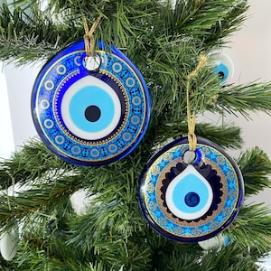 Puede incluir: Dos adornos azules y dorados de ojo turco colgando de un árbol de Navidad artificial verde. Los adornos son redondos y tienen un diseño de ojo blanco y azul con detalles dorados.