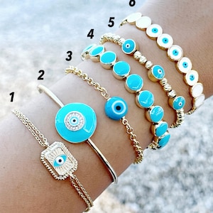 Könnte beinhalten: Ein Set aus sechs goldenen Armbändern mit blauen bösen Augen-Anhängern. Die Armbänder sind auf dem Handgelenk einer Person übereinander gelegt. Die Armbänder bestehen aus verschiedenen Materialien, darunter Perlen, Ketten und Metall.