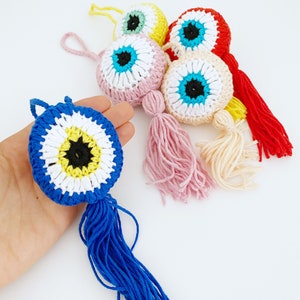 May include: Five crocheted evil eye charms with tassels in various colors, including pink, blue, yellow, red, and cream. The charms are all crocheted in a similar pattern, with a blue eye in the center and a white circle around it. The tassels are all different colors and lengths.