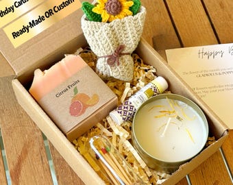 May include: A gift box with a crochet sunflower bouquet, a bar of soap labelled "Citrus Fruits", a candle, lip balm, matches, and a birthday card. The card says "Happy Birthday". The box is filled with shredded paper.