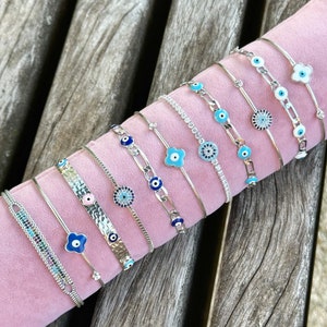 May include: A collection of silver-toned bracelets with various designs, including evil eye and floral motifs. The bracelets are displayed on a pink surface, showcasing a range of styles with blue, white, and clear accents. The bracelets are made of metal.