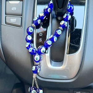 Op de afbeelding: Een blauwe auto-hanger met kralen, elk met een evil eye-ontwerp. De hanger hangt voor de versnellingspook van een auto.