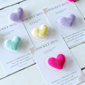 Può includere: Una collezione di Pocket Hugs fatti a mano a forma di cuore all'uncinetto, in vari colori, tra cui viola, giallo, turchese e rosa. Ogni cuore è posto su una carta con il testo "POCKET HUG" e un messaggio sull'amore e gli abbracci.