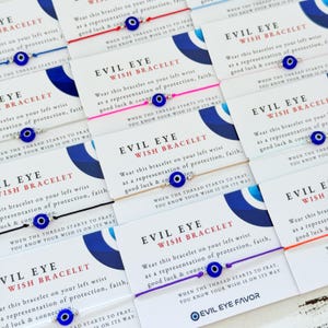 Puede incluir: Una colección de pulseras de deseos de Evil Eye en tarjetas. Cada pulsera presenta un dije de ojo azul y está ensartada en un cordón de color. Las tarjetas incluyen texto sobre protección y buena suerte.