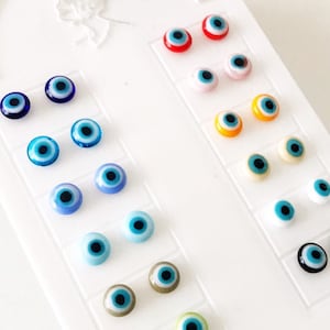 Pode incluir: Um cartão branco com o texto "FASHION EARRINGS" impresso nele. O cartão tem uma exibição de 20 pares de pequenos brincos de olho turco coloridos.