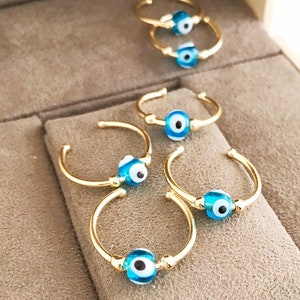 May include: Gold-toned rings with a blue and white evil eye bead. The rings are displayed inside a gray jewelry box. The evil eye bead is a popular symbol believed to offer protection against negative energy. The rings are delicate and stylish.