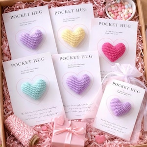 Può includere: Una scatola regalo piena di biglietti con la scritta "Pocket Hug" e cuori all'uncinetto in vari colori: viola, giallo, rosa e turchese. La scatola è decorata con nastro rosa e carta, con una piccola scatola regalo e una bobina di spago rosa.