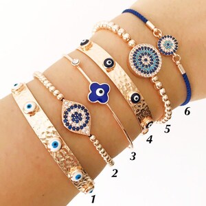 May include: A set of seven gold bracelets with blue evil eye charms. The bracelets are stacked on a person's wrist. The bracelets are numbered 1 through 6.