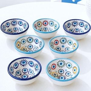 May include: A set of six small ceramic bowls with a white base and hand-painted evil eye motifs in blue, green, yellow, and red. Each bowl features a circular pattern of concentric rings and dots, with a solid colored rim in either blue or light blue.