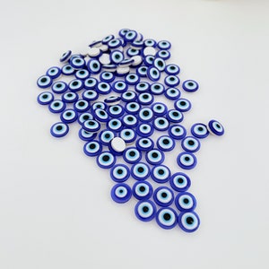 May include: A large group of blue evil eye charms with white centers and black pupils. The charms are scattered on a white surface.