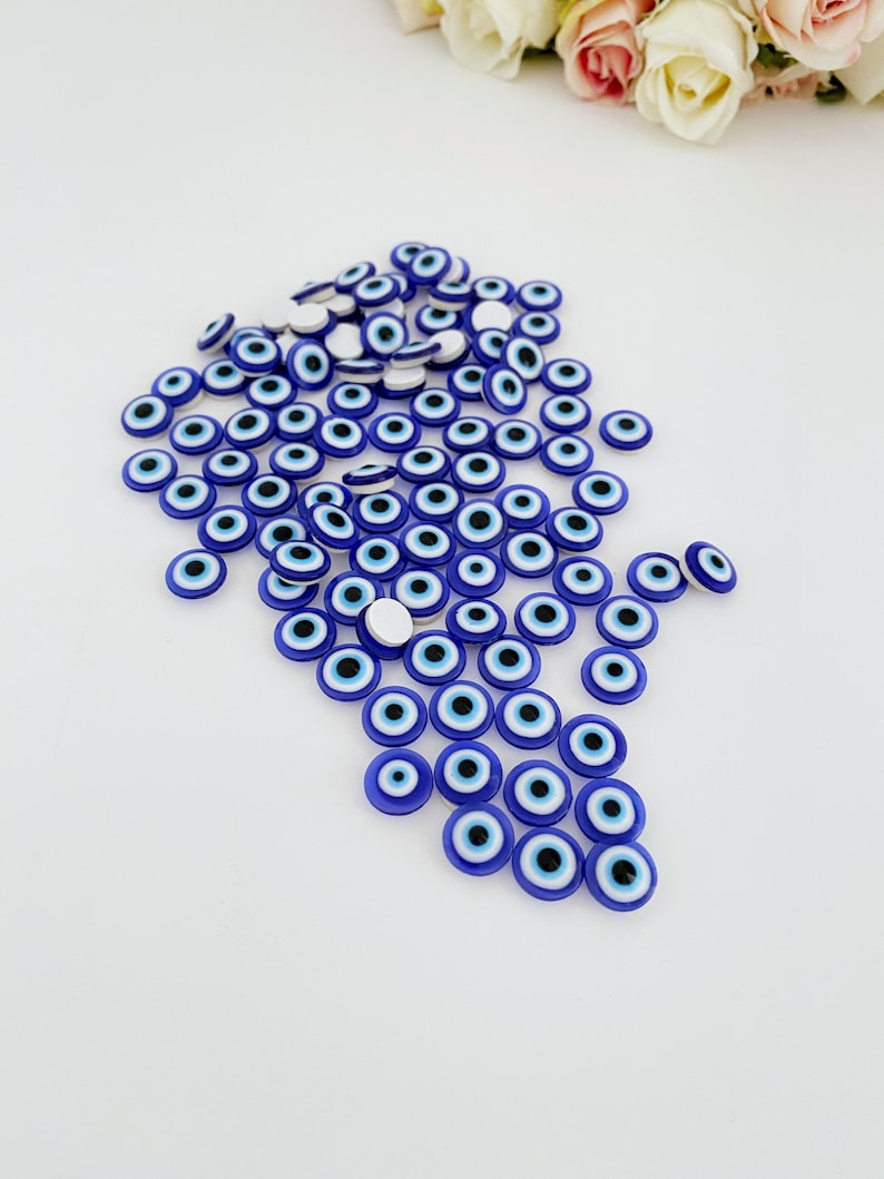 May include: A large group of blue evil eye charms with white centers and black pupils. The charms are scattered on a white surface.