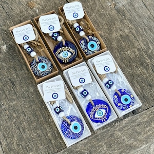 May include: Six blue and white evil eye charms with white beads and twine hanging from boxes. The boxes have white paper with the text "The Evil Eye" and a smaller evil eye charm.  The text reads "Stares back at the world to shield you from negativity and harm."