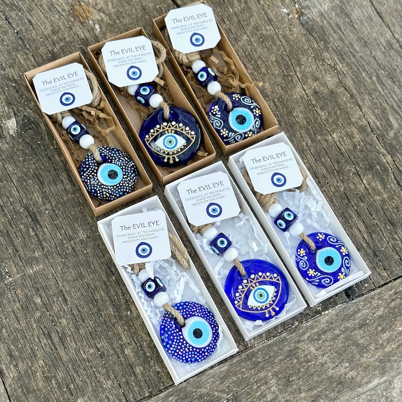 May include: Six blue and white evil eye charms with white beads and twine hanging from boxes. The boxes have white paper with the text "The Evil Eye" and a smaller evil eye charm.  The text reads "Stares back at the world to shield you from negativity and harm."