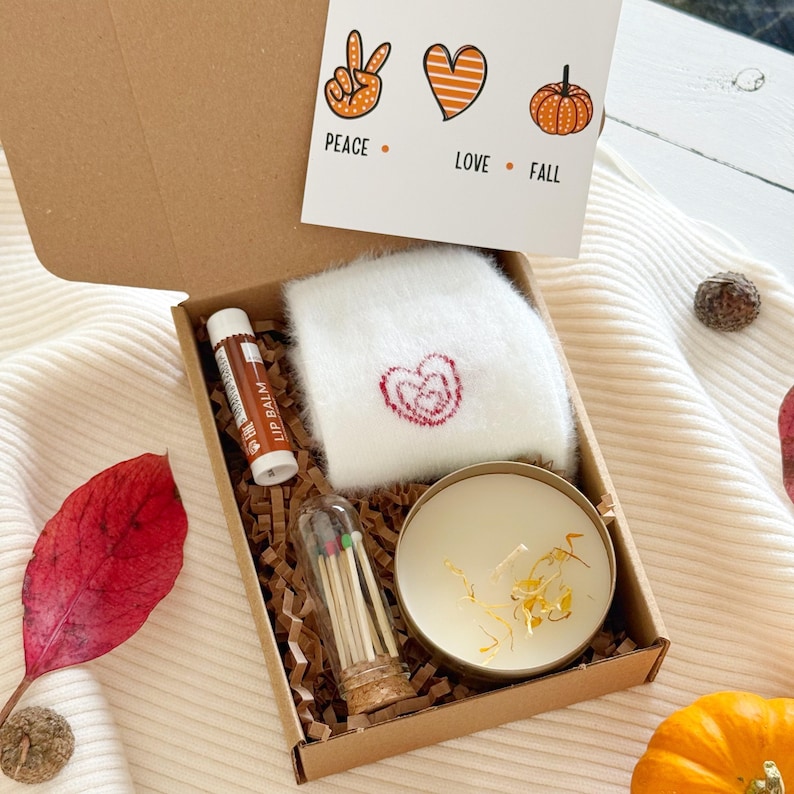 May include: A cardboard gift box containing a pair of white fuzzy socks with a red heart, a brown lip balm tube, a glass vial of matches with colored tips, and a round candle with dried petals. A card reads PEACE, LOVE, FALL with icons.