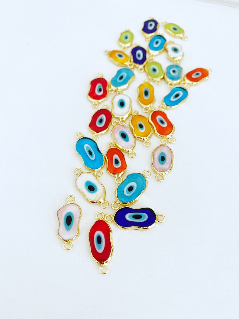 May include: A collection of colorful evil eye charms with gold accents. The charms are shaped like an eye and have a blue pupil. They are arranged in a pile on a white surface.