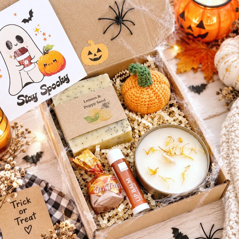 May include: A cardboard gift box containing a bar of lemon and poppy seed soap, a crocheted orange pumpkin, a white candle in a gold tin, a brown lip balm tube, and a wrapped candy. A card with a ghost and the text Stay Spooky is included.
