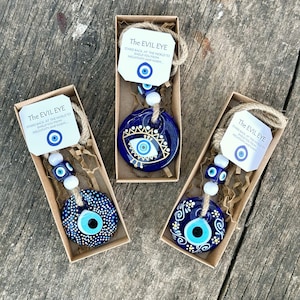 May include: Three small blue and white evil eye charms in gift boxes. Each charm has a different design and is strung on a twine with white beads. The boxes have a small tag with the text "The Evil Eye" and a description of the charm.