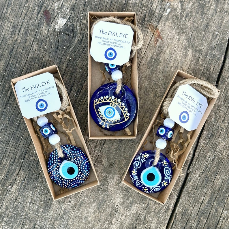 May include: Three small blue and white evil eye charms in gift boxes. Each charm has a different design and is strung on a twine with white beads. The boxes have a small tag with the text "The Evil Eye" and a description of the charm.