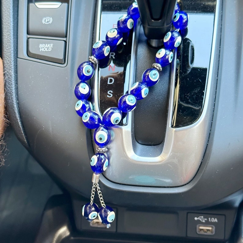 Op de afbeelding: Een blauwe auto-hanger met kralen, elk met een evil eye-ontwerp. De hanger hangt voor de versnellingspook van een auto.
