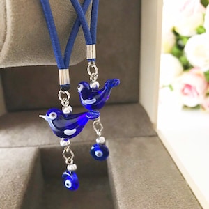 May include: Two blue glass bird charms with white accents hanging from a blue cord with silver beads and blue evil eye charms.