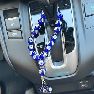 Op de afbeelding: Een blauwe kralen auto-hanger met meerdere glazen kralen, elk met een wit en blauw oogontwerp. De hanger hangt in een auto, met een zilveren ketting en een kwastje van kleinere oogkralen.